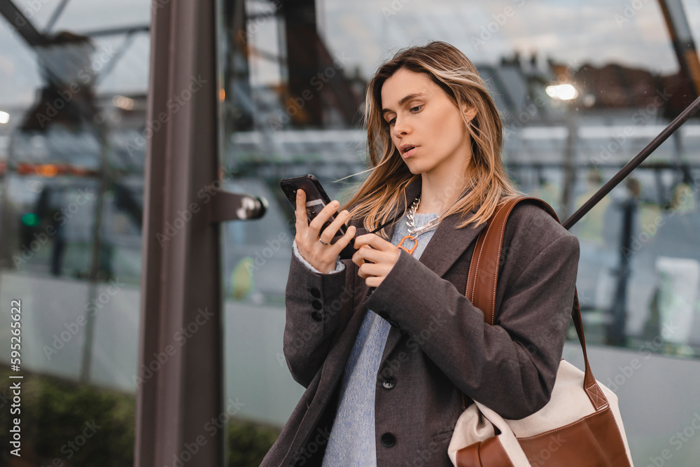 Fototapeta premium Attractive blonde woman standing on bus station and wait for a bus, using mobile phone, read sms, text, news. Portrait of calm confident female blogger using cellphone reading.