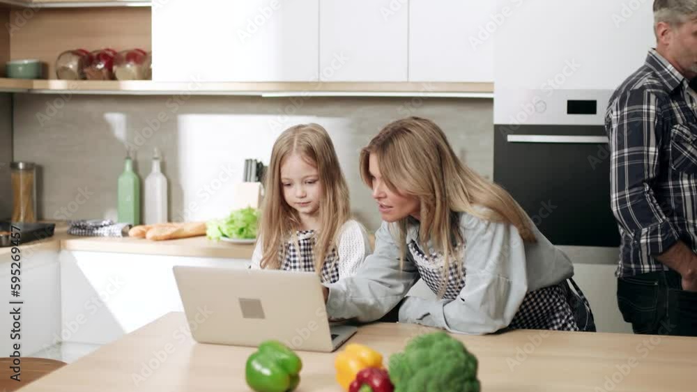 Smiling blond mother help her cute little daughter with homework on laptop computer at distance remote education while busy father has working business call by cellphone at domestic kitchen at home Smiling blond mother help her cute little daughter with homework on laptop computer at distance remote education while busy father has working business call by cellphone at domestic kitchen at home