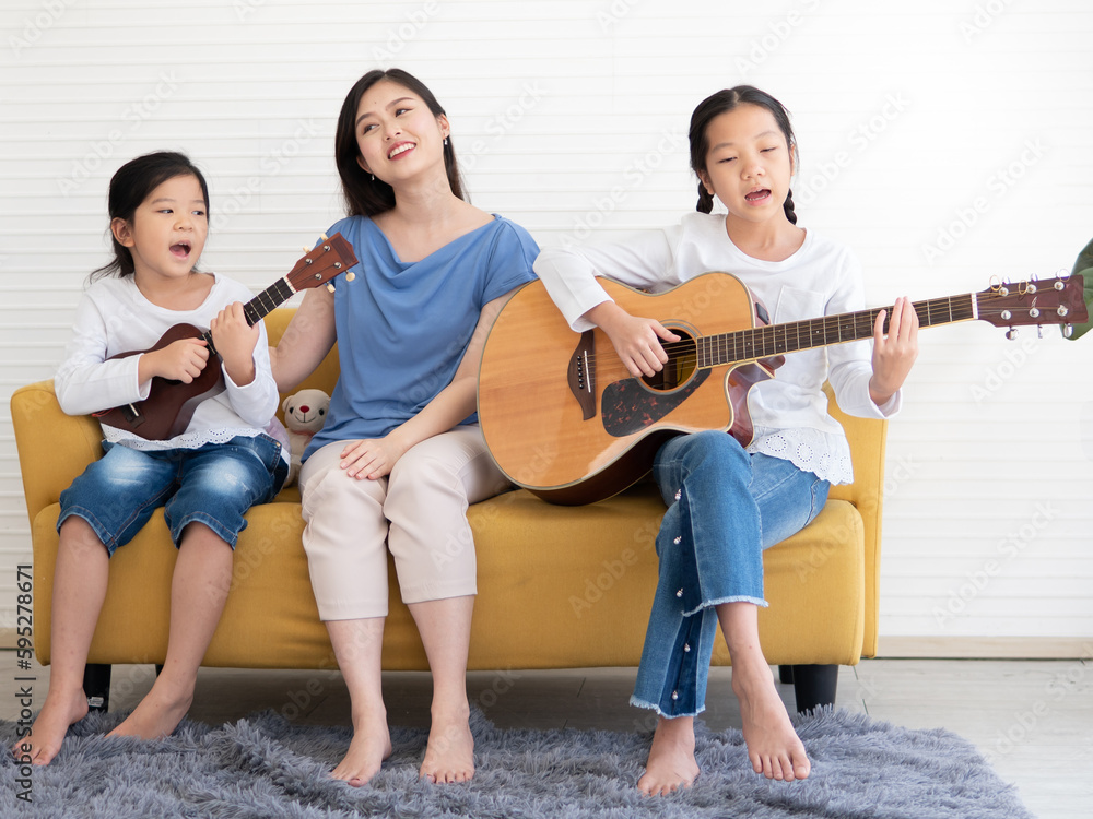 Obraz premium Pretty little girl sitting at couch enjoy playing acoustic guitar with her mother and sibling. Adorable kid playing ukulele with her sister and mom. Asian family guitarist singing together at leisure.