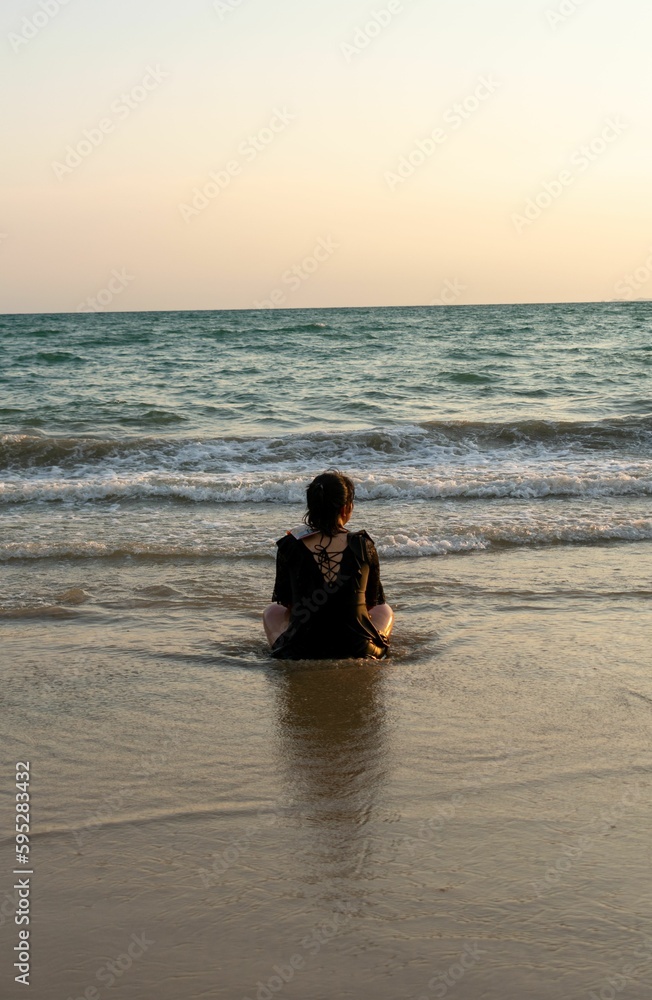 Woman sitting on a sandy shore of a beach, looking as the sun sets in the distance