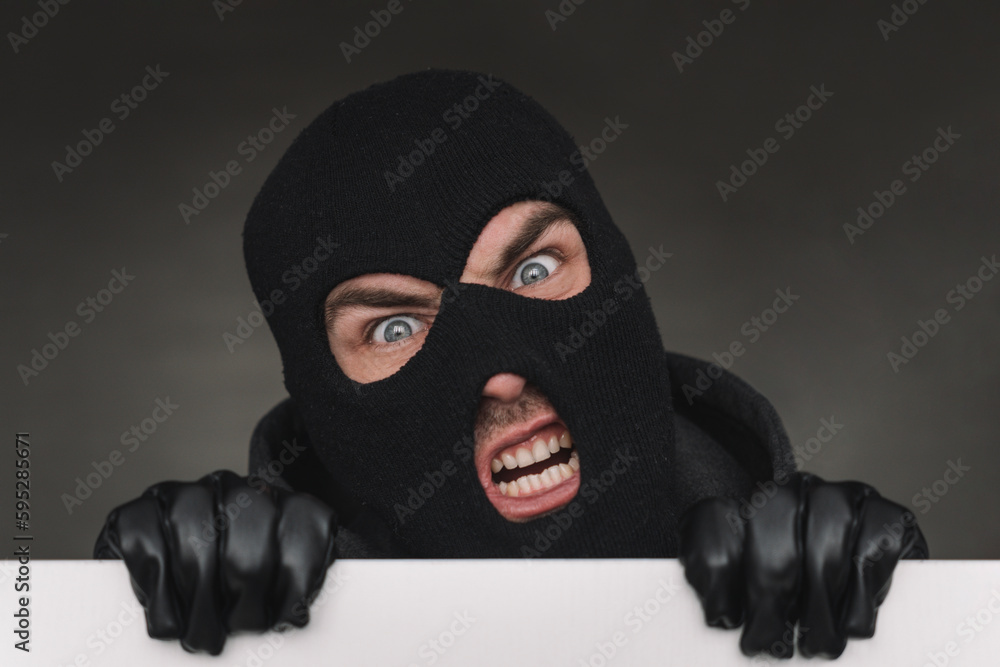 An evil thief in a mask looks out from behind a white board. evil bandit with a mask on his face. An evil thief in a mask looks out from behind a white board. evil bandit with a mask on his face.