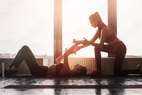 Two young girls train together in modern gym on background of panoramic windows. Young teen girl is doing exercises with dumbbells lying on floor on back.