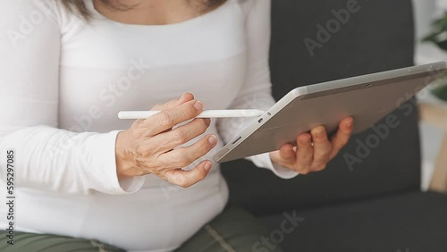 From above of positive adult female in casual outfit with headphones and tablet sitting on sofa with pillows in light studio photo