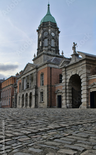 Evening at Dublin Castle. Cubic stone pavement, a medieval fortress, brick walls, a clock tower, and a green dome against a background of contrails in the blue sky