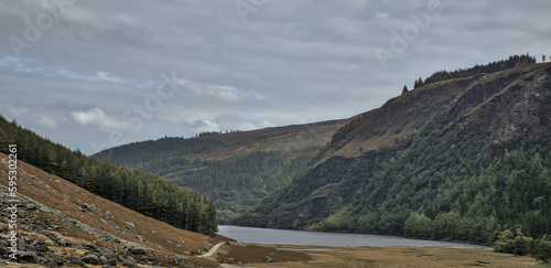 Through a barren meadow, a hiking path leads to a lake encircled by pine trees and lush rocks. Ireland's Wicklow Mountains