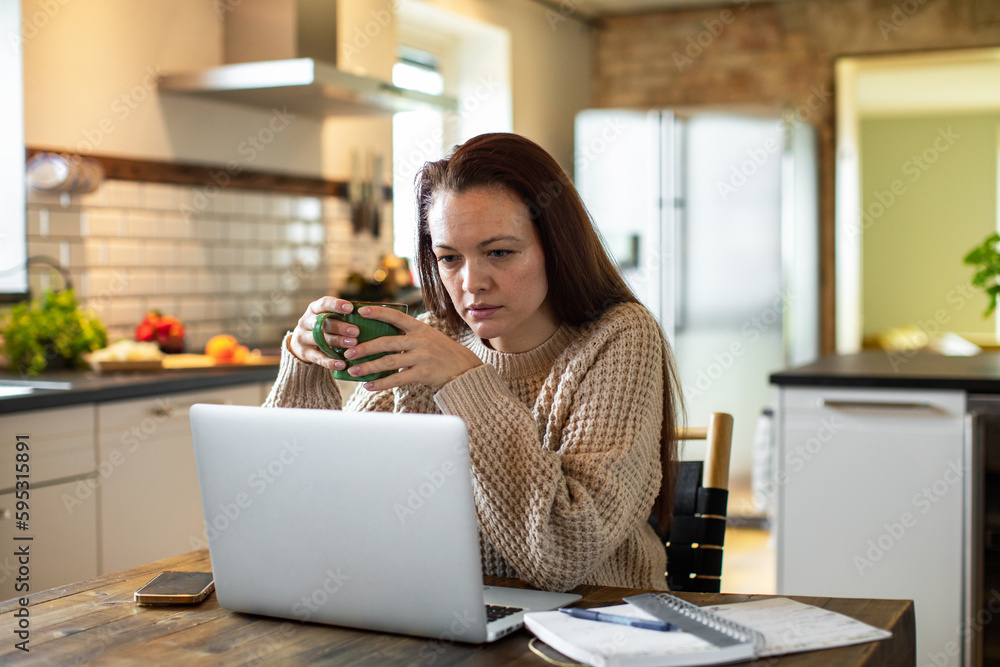 Young adult woman using a laptop in a kitchen in the morning Young adult woman using a laptop in a kitchen in the morning