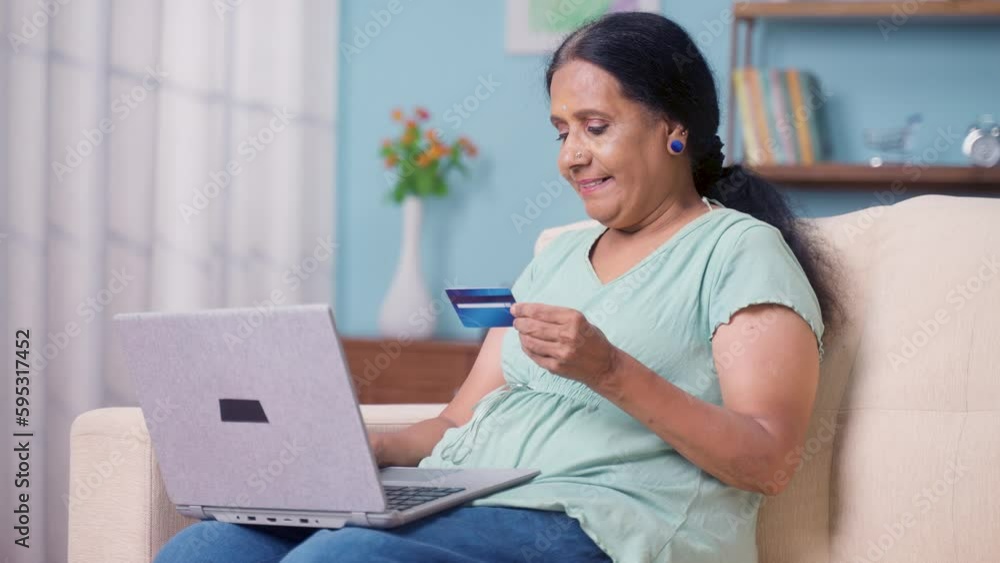 Happy Indian elderly senior woman doing online shopping by using credit card on laptop while sitting on sofa at home - concept of bill payment, e commerce and financial or banking. Happy Indian elderly senior woman doing online shopping by using credit card on laptop while sitting on sofa at home - concept of bill payment, e commerce and financial or banking.