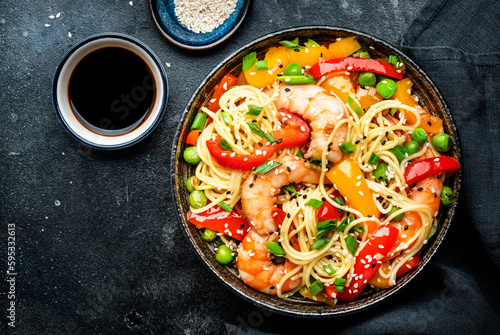 Stir fry egg noodles with shrimps, paprika, green pea, chives and sesame seeds bowl. Asian cuisine dish. Black kitchen table background, top view