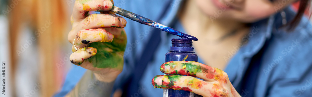 Smiling young woman in apron holding blue paint jar and a paintbrush, working on a painting in workshop Smiling young woman in apron holding blue paint jar and a paintbrush, working on a painting in workshop