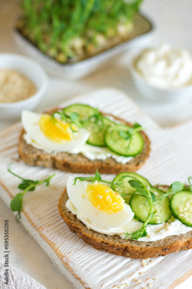 Cottage cheese sandwiches on whole grain bread with half an egg and cucumber with microgreens on a wooden board, the background is blurred