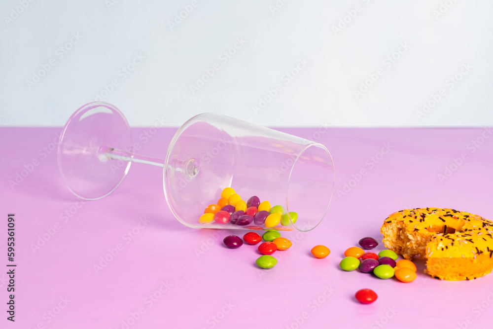 Fallen glass cup with a colorful round glazed candies and a bitten donut. isolated on pastel purple background.