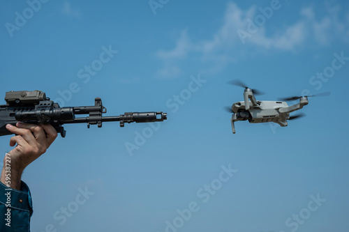 A man aims to shoot a rifle at a flying drone against a blue sky. 