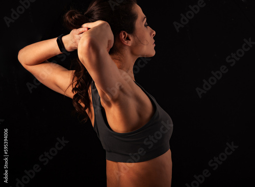 Sporty muscular woman with ponytail doing stretching workout of the shoulders, blades in sport bra, holding hands behind the head on dark grey background with empty space.