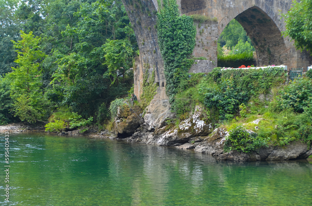 A fisherman rests from fishing, just below the Roman bridge in Cangas de Onis, in Asturias, Spain, below, the Sella river, surrounded by trees and green vegetation.