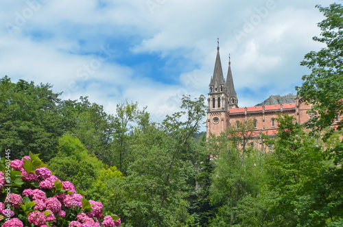 The Sanctuary of Covadonga in the distance  surrounded by green trees and flowers with a sunny sky and clouds.