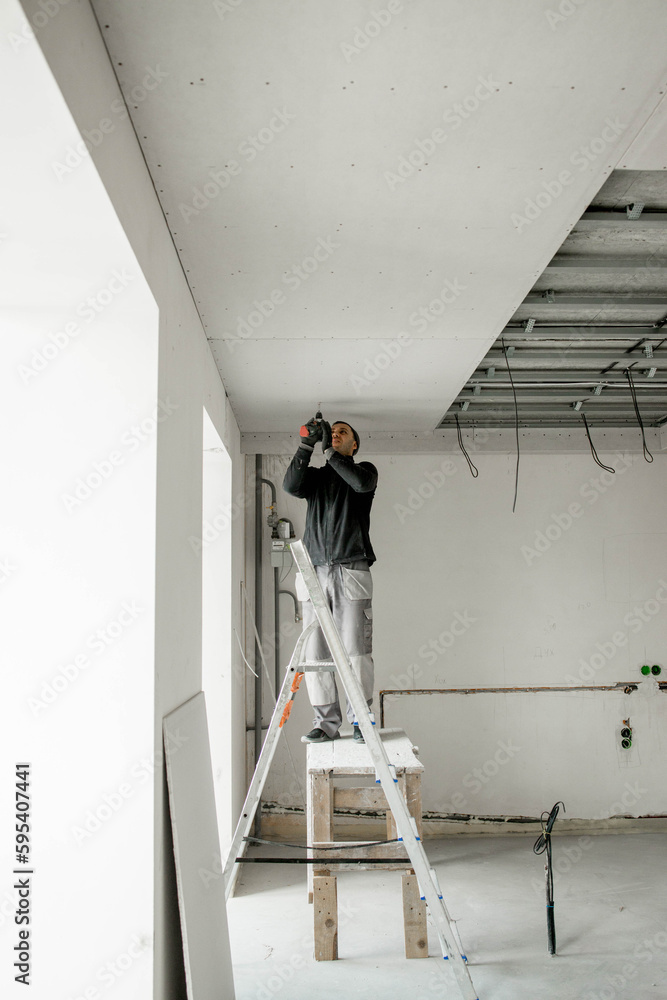 Construction worker assemble a suspended ceiling with drywall and fixing the drywall to the ceiling metal frame with screwdriver. Renovation, construction and do it yourself DIY concept. Construction worker assemble a suspended ceiling with drywall and fixing the drywall to the ceiling metal frame with screwdriver. Renovation, construction and do it yourself DIY concept.