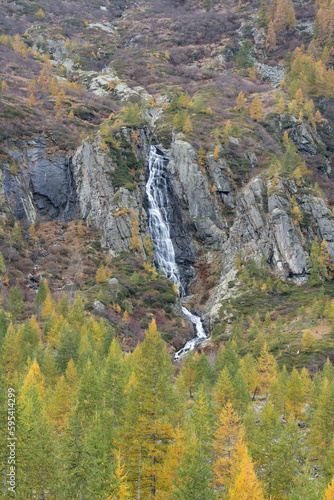 mountain forest in the valley, yellow orange colored trees and waterfall over a rocky cliff in autumn