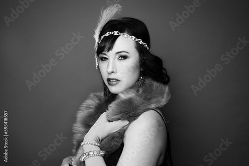 Black and white studio portrait of a caucasian woman wearing 1920s attire. She is wearing a fringe trimmed dress, fur scarf, feathered headpiece and gloves. 