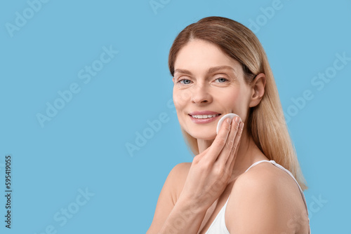Beautiful woman removing makeup with cotton pad on light blue background, space for text