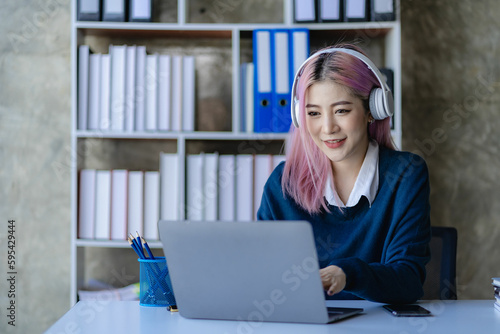 Asian female college student wearing headphones studying online with laptop computer surfing internet meeting Studying online, smiling happily at home.