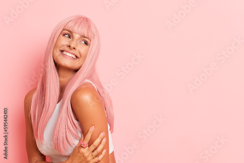 Pretty girl with long pink hair and beaming smile posing aside of studio background, holds one hand on her left arm, looks at free space for your advertising, happy life concept, copy space, high