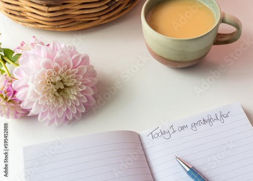 High angle closeup of notebook page with "Today I am grateful for" written on it, next to pink dahlia and cup of tea (selective focus)
