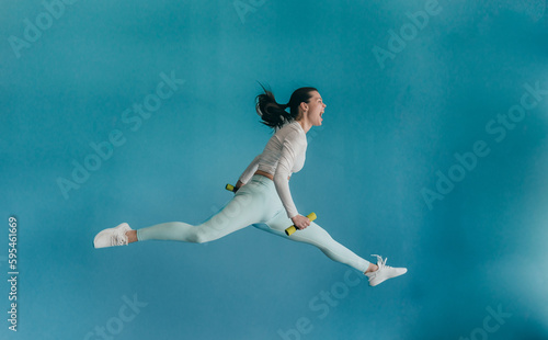 Young brunette American woman in sportswear jumps high holds dumbbells screams against blue wall. Healthy people. Fit American girl running shouting out loud. Sport club, weight loss concept.