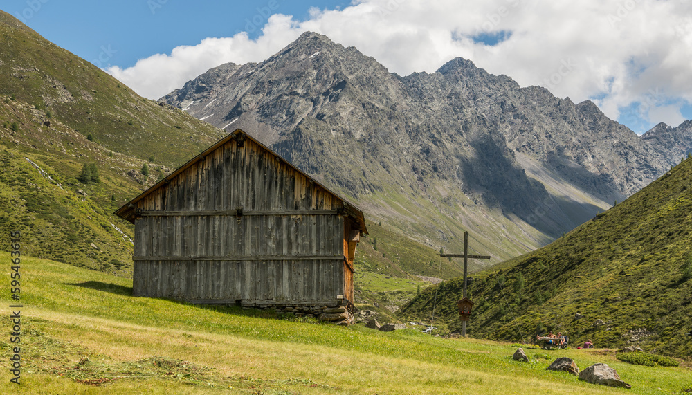 Summer alpine view of a wooden barn in a fresh alpine meadow  barn and a wooden cross. Mountains in the background. Umhausen, Austria