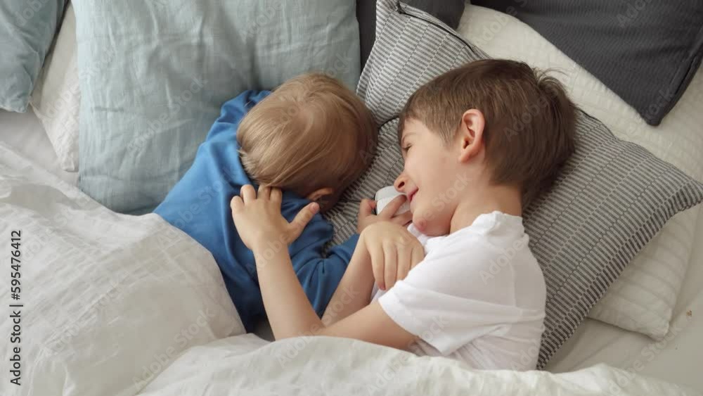 Two cute boys lying in bed on soft cushions and smiling at each other. Concept of happy childhood, fun at home and cheerful kids Two cute boys lying in bed on soft cushions and smiling at each other. Concept of happy childhood, fun at home and cheerful kids