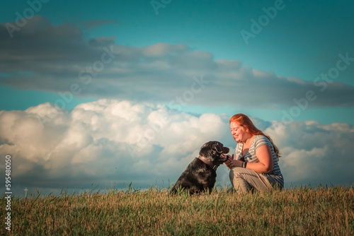 Portrait of a young beautiful plump red-haired girl with a black labrador in a field against the sky. © shymar27