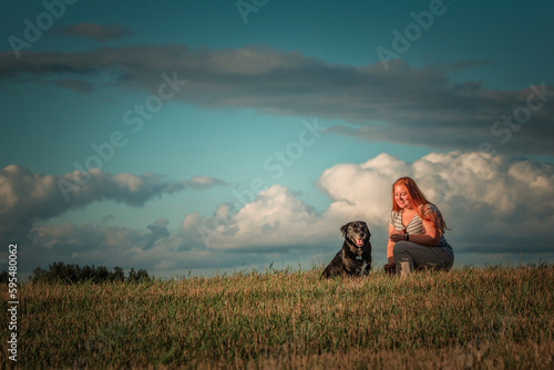 Portrait of a young beautiful plump red-haired girl with a black labrador in a field against the sky.