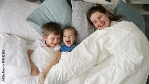 Happy laughing family with two kids hiding under blanket in bed and throwing it off. Concept of family happiness, relaxing at home, having fun in bed, parent and cheerful kids.