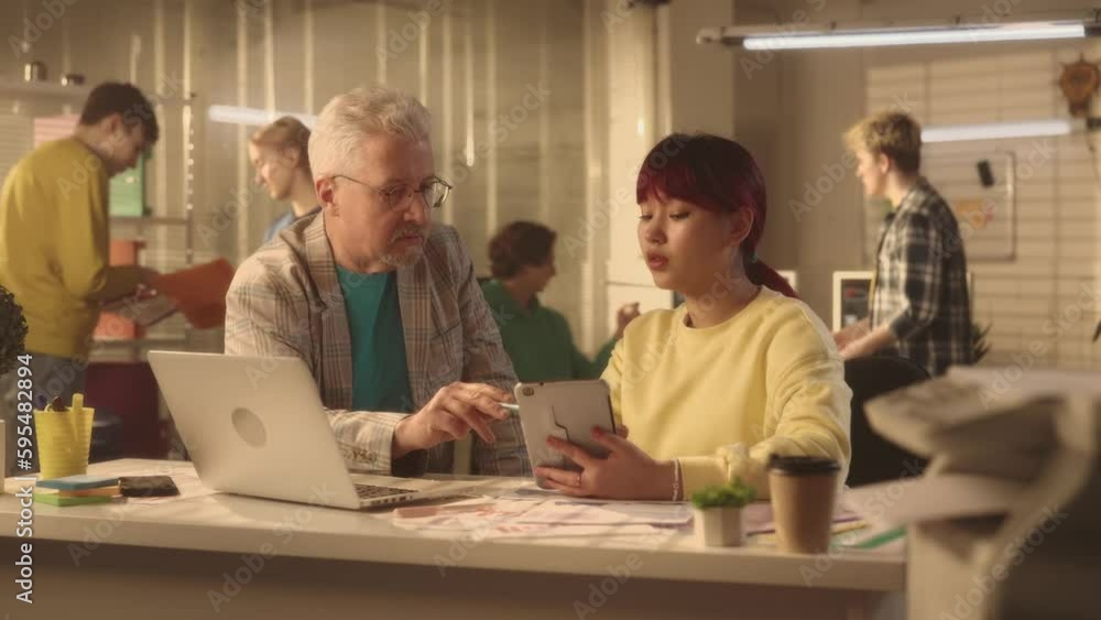 Aged man is discussing a new project with young asian woman on a laptop and tablet. A businessman and manager works in an office. A young creative team of managers are working in the background. Aged man is discussing a new project with young asian woman on a laptop and tablet. A businessman and manager works in an office. A young creative team of managers are working in the background.