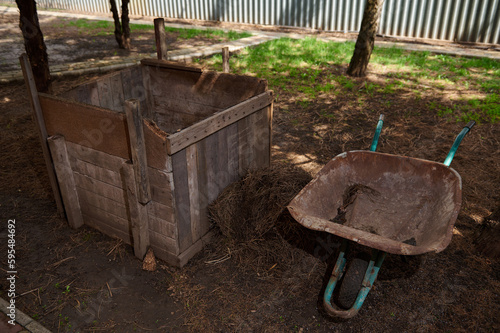 Still life with a wooden compost bin and wheelbarrow. Close up DIY compost bin to improve the fertility and soil structure in the garden. Zero waste concept. Organic farming. Sustainability