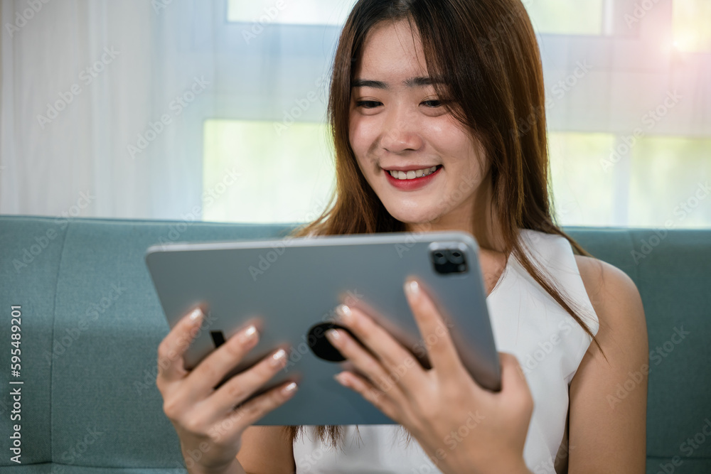 Happy Asian woman playing smart tablet at home while relaxing on sofa living room, Beautiful female sitting on couch using digital tablet pc, technology social media Happy Asian woman playing smart tablet at home while relaxing on sofa living room, Beautiful female sitting on couch using digital tablet pc, technology social media