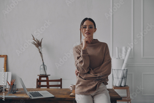 Confident young woman looking at camera while leaning at the desk in creative office