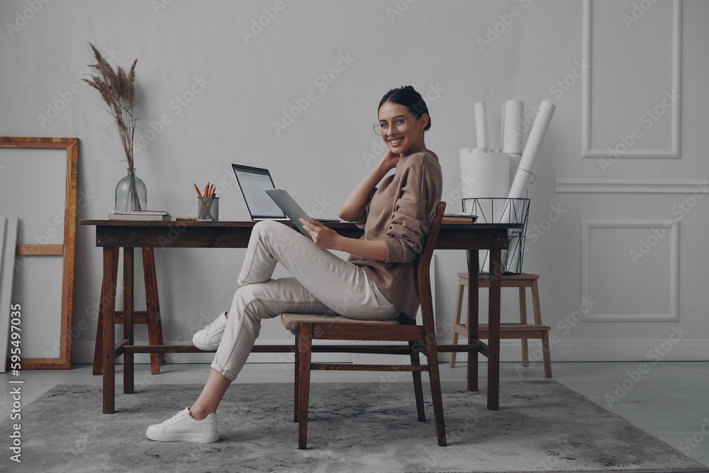 Beautiful young woman holding digital tablet and smiling while sitting at her workplace