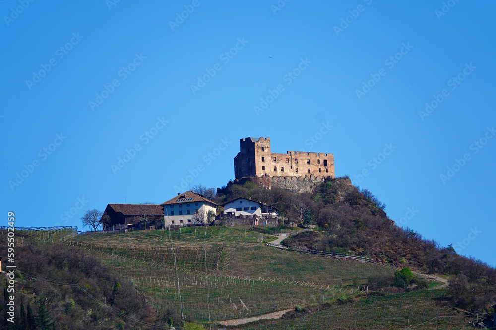 Eine freistehende Burg auf einem Hügel in Südtirol mit einigen unterhalb liegenden Häusern, vor strahlend blauem Himmel