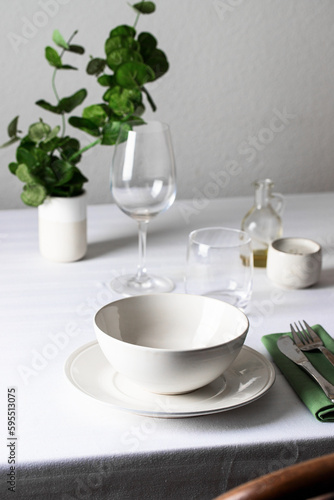 Table setting. Ceramic bowl and plate, silver cutlery, linen napkin, water and wine galss , eucalyptus branch on white table background. Restaurante place setting