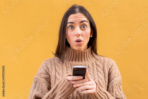 Young brunette woman at outdoors looking at the camera while using the mobile with surprised expression