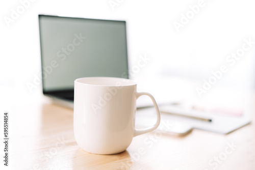 White mug without inscriptions in front of a laptop and a notepad on a wooden table.