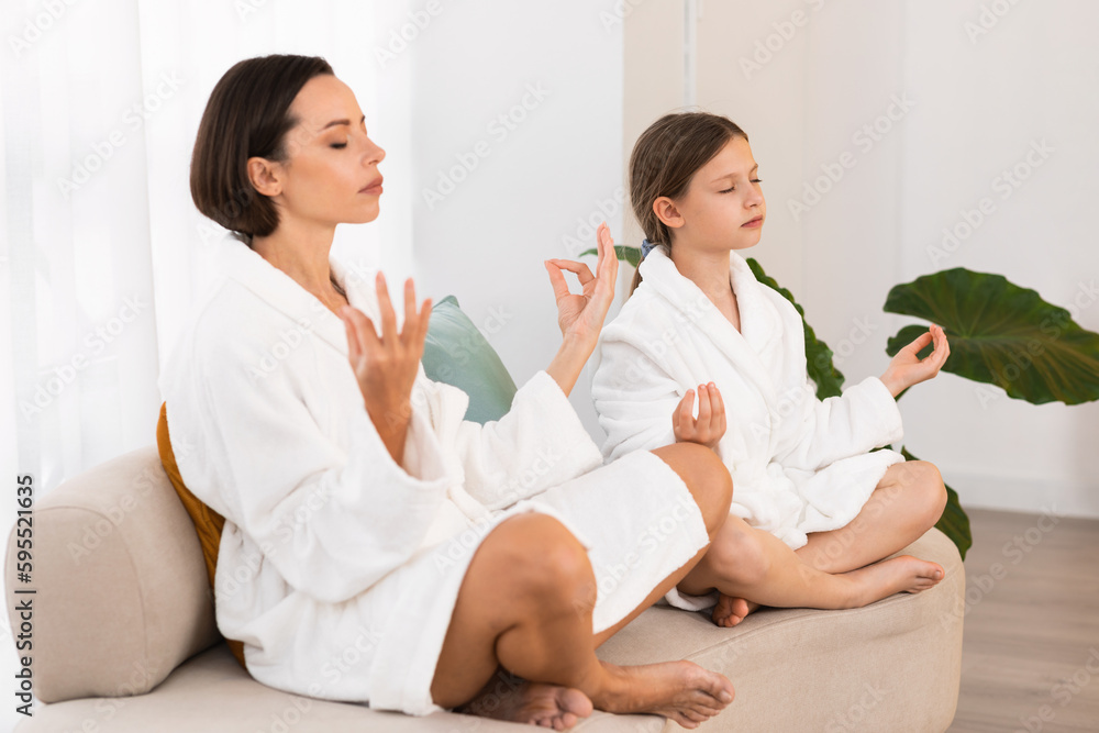 Fototapeta premium Zen. Peaceful Young Mom And Daughter In Bathrobes Meditating Together At Home