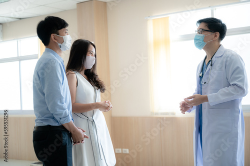 Young male doctor wearing covid-19 safety mask in conversation with young couple in clinic and hospital passage