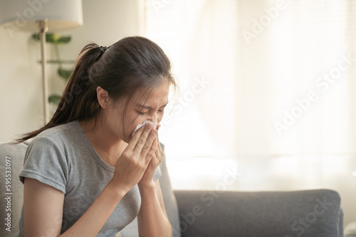 Asian woman with a cold blowing her nose at home on the couch, temperature and allergy