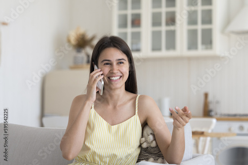 Cheerful Hispanic late teen girl speaking on cellphone, sitting on home couch, chatting, smiling, laughing. Happy pretty young woman enjoying conversation on mobile phone
