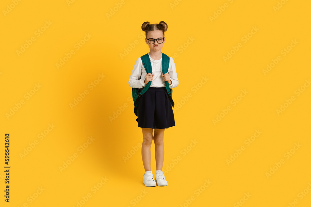 Portrait Of Grumpy Little Schoolgirl With Backpack Standing Over Yellow Studio Background Portrait Of Grumpy Little Schoolgirl With Backpack Standing Over Yellow Studio Background