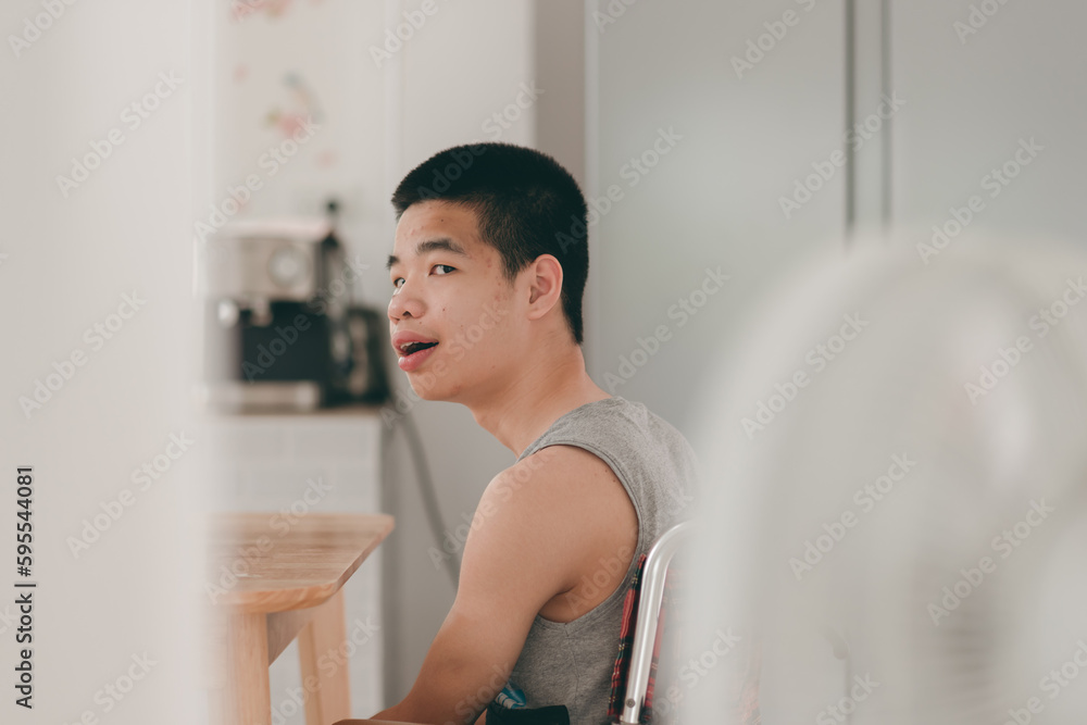 Young man with disability wear a tank top to cool off in the summer. Sit on wheelchair using laptop with a fan in the house,hospital,school,nursery, Taking care of health to suit the heat air. Young man with disability wear a tank top to cool off in the summer. Sit on wheelchair using laptop with a fan in the house,hospital,school,nursery, Taking care of health to suit the heat air.