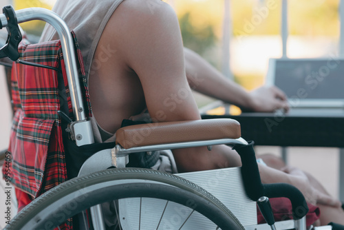 Young man with disability wear a tank top to cool off in the summer. Sit on wheelchair using laptop with a fan in the house,hospital,school,nursery, Taking care of health to suit the heat air.