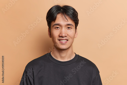 Portrait of handsome young Vietnamese man smiles gladfully shows white teeth has joyful expression dressed in black t shirt rejoices having meeting with friend isolated over brown background