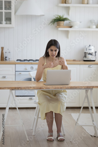 Serious engaged young adult Latin girl talking on video call on laptop, speaking at screen, display, using wireless earphone in ear for wireless connection, working at kitchen table